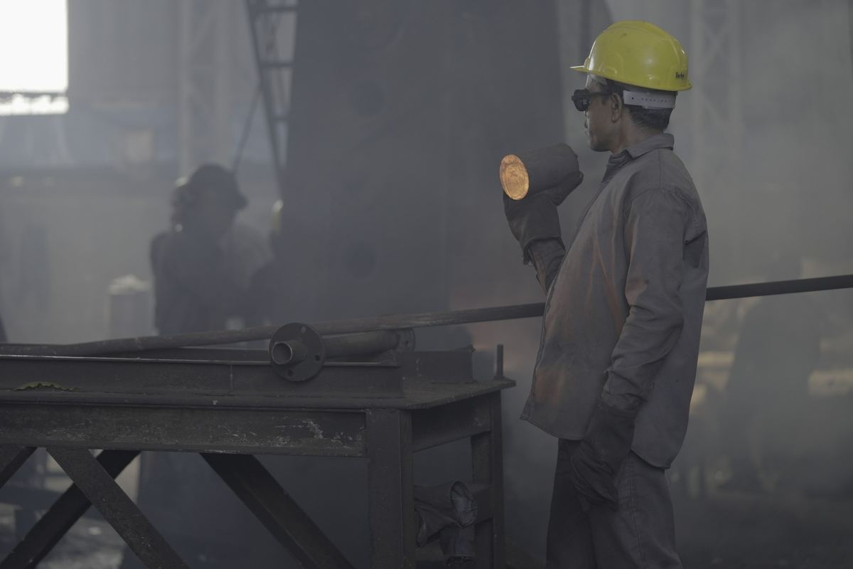 Forging shop worker in safety gear holding a hot billet with tongs